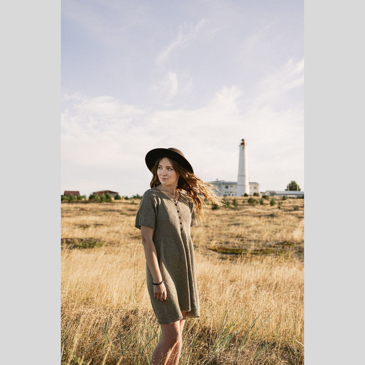 Woman in a knitted dress standing in a field with a lighthouse in the background. This is a page from Laine Book "Unfolding Landscapes"
