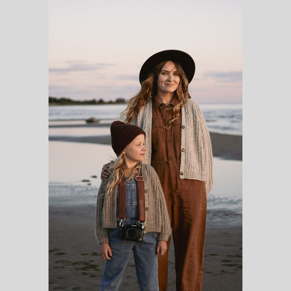 Two people standing on a beach at sunset, wearing hats and cardigans. This is a page from Laine Book "Unfolding Landscapes"