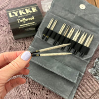 Set of LYKKE Driftwood knitting needles in a gray case on a textured surface. Hand holding needles above set.