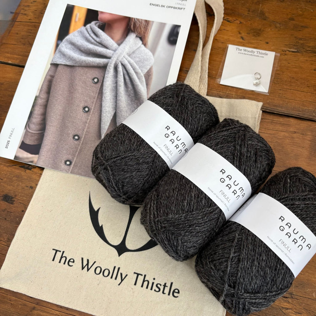 Magdalenasjal Shawl Kit from The Woolly Thistle including Rauma Finullgarn yarn in dark grey, printed pattern, stitch marker, and branded tote bag shown at an angle