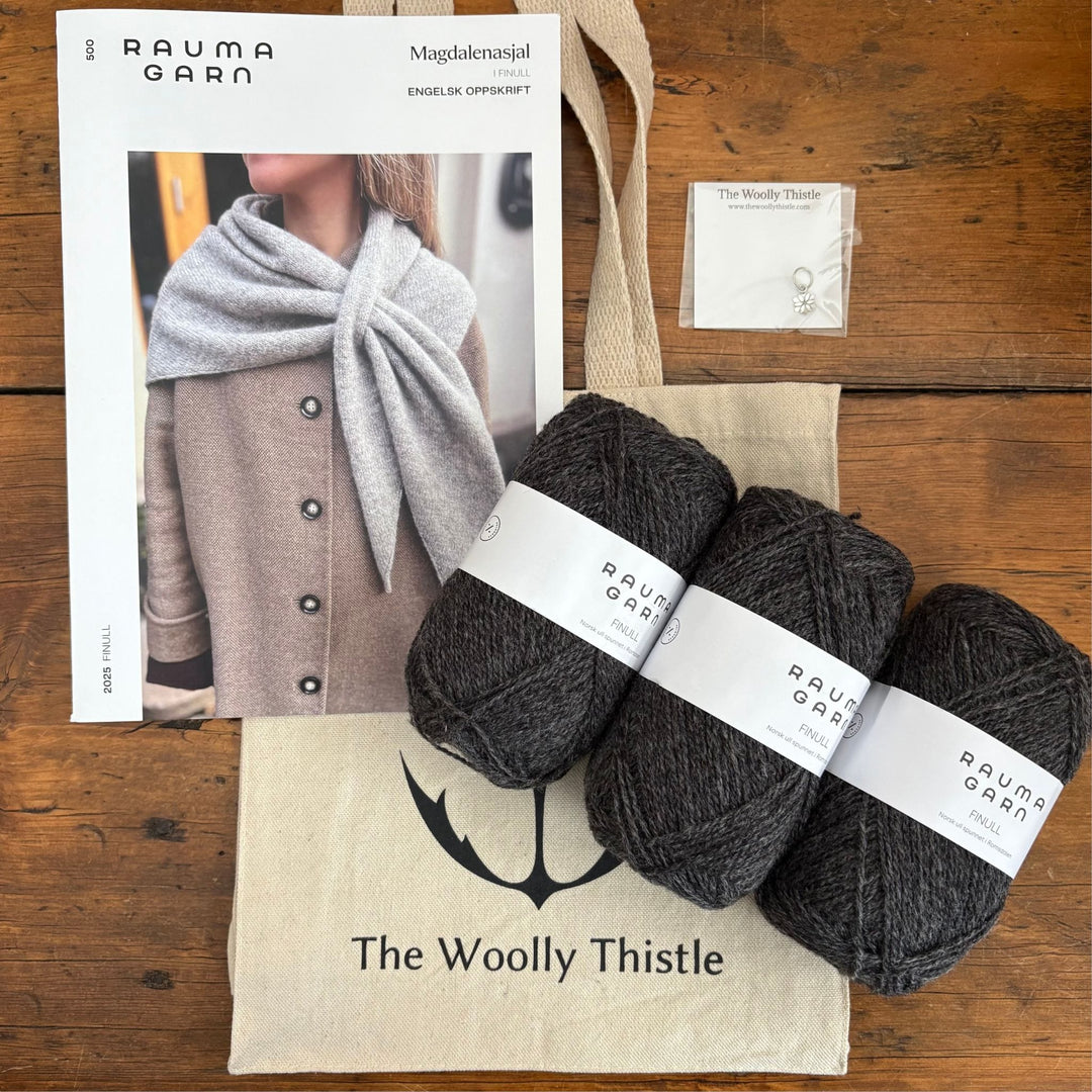 Magdalenasjal Shawl Kit components shown on a wooden surface including three balls of Rauma Finullgarn in dark grey, printed pattern, stitch marker, and The Woolly Thistle tote bag.