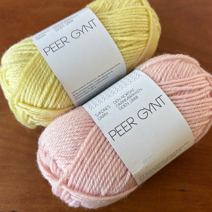 Two skeins of yarn, one yellow and one pink, with 'Peer Gynt' labels on a wooden surface.