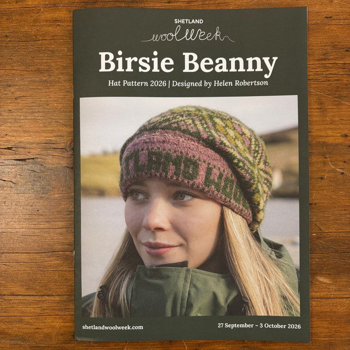 Shetland Wool Week 2026 Birsie Beanny hat pattern booklet on a wooden surface. Cover shows a woman wearing a handknit colorwork hat.