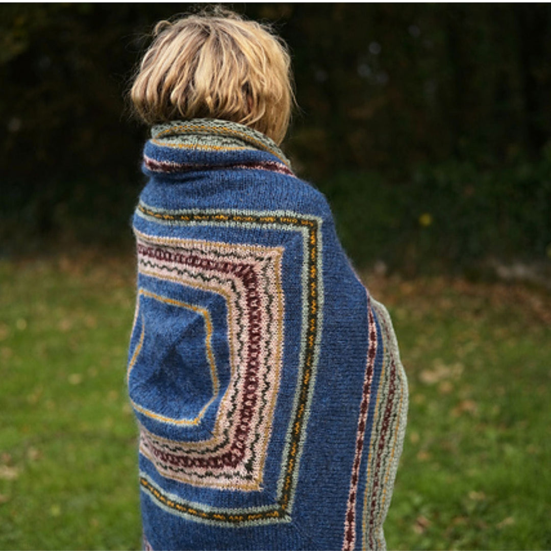 Side view of person wearing a blue knitted shawl with colorful patterns, standing outdoors. Shawl design is Moon Tide Shawl. 