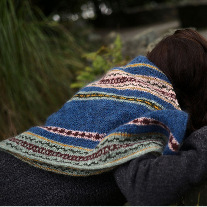 Side view of Person wearing a blue knitted shawl with colorful patterns, sitting outdoors. Shawl design is Moon Tide Shawl. 