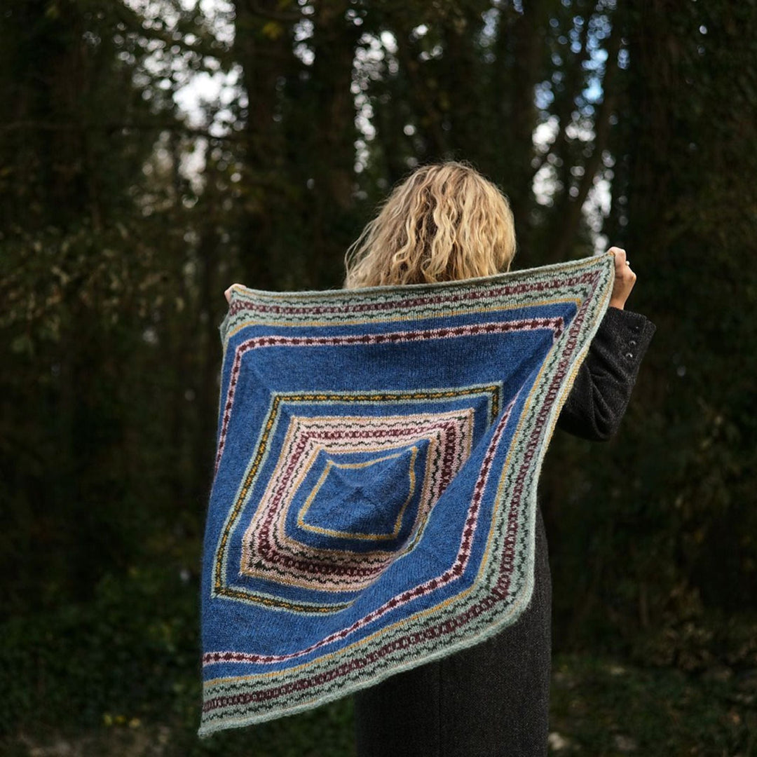 Back view of Person holding a knitted, colorful shawl outdoors with trees in the background. Shawl design is Mood Tide Shawl.
