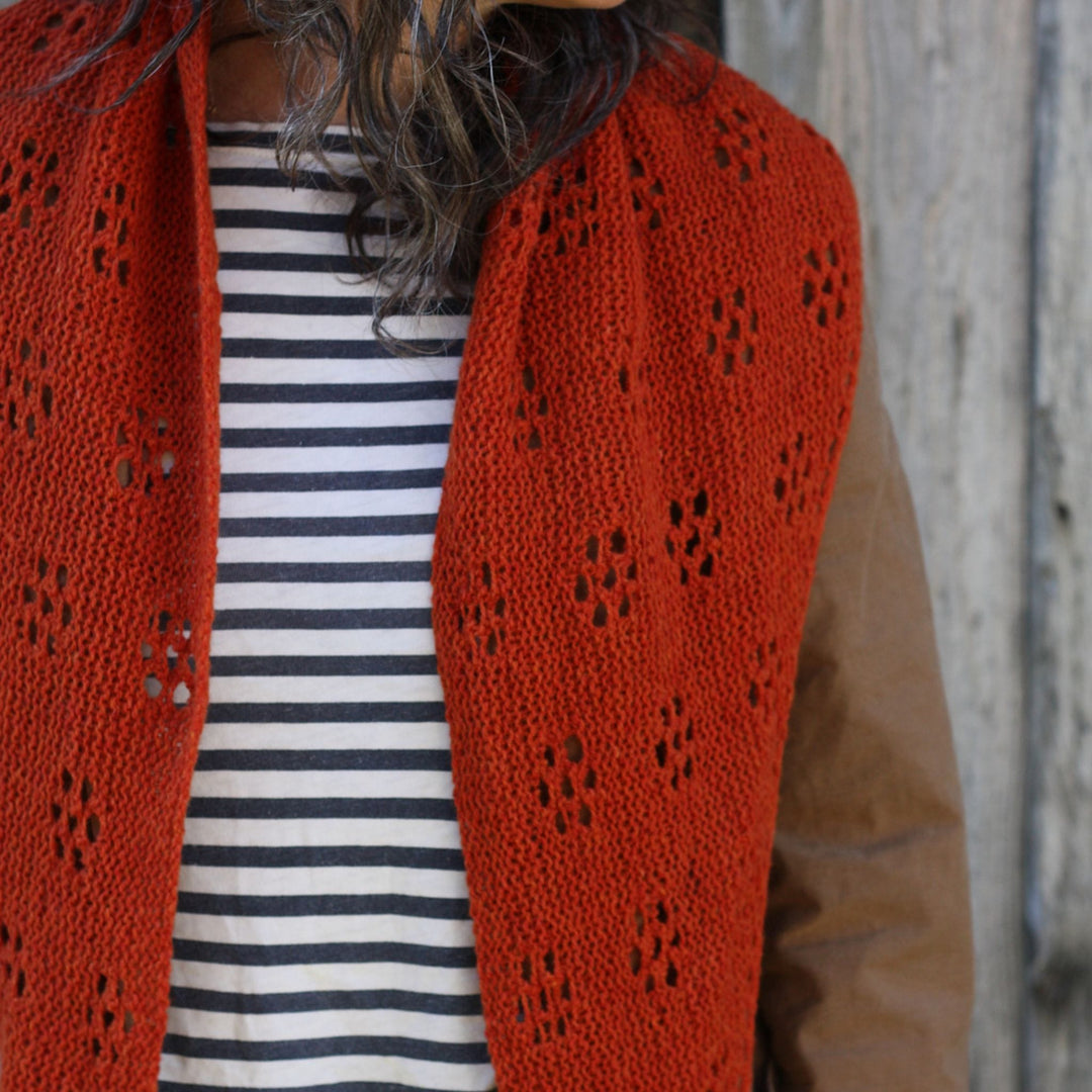 Person wearing a red knitted shawl over a striped shirt with a blurred wooden background. Shawl design shown is Ghost Flowers Shawl by Amy Christoffers. 