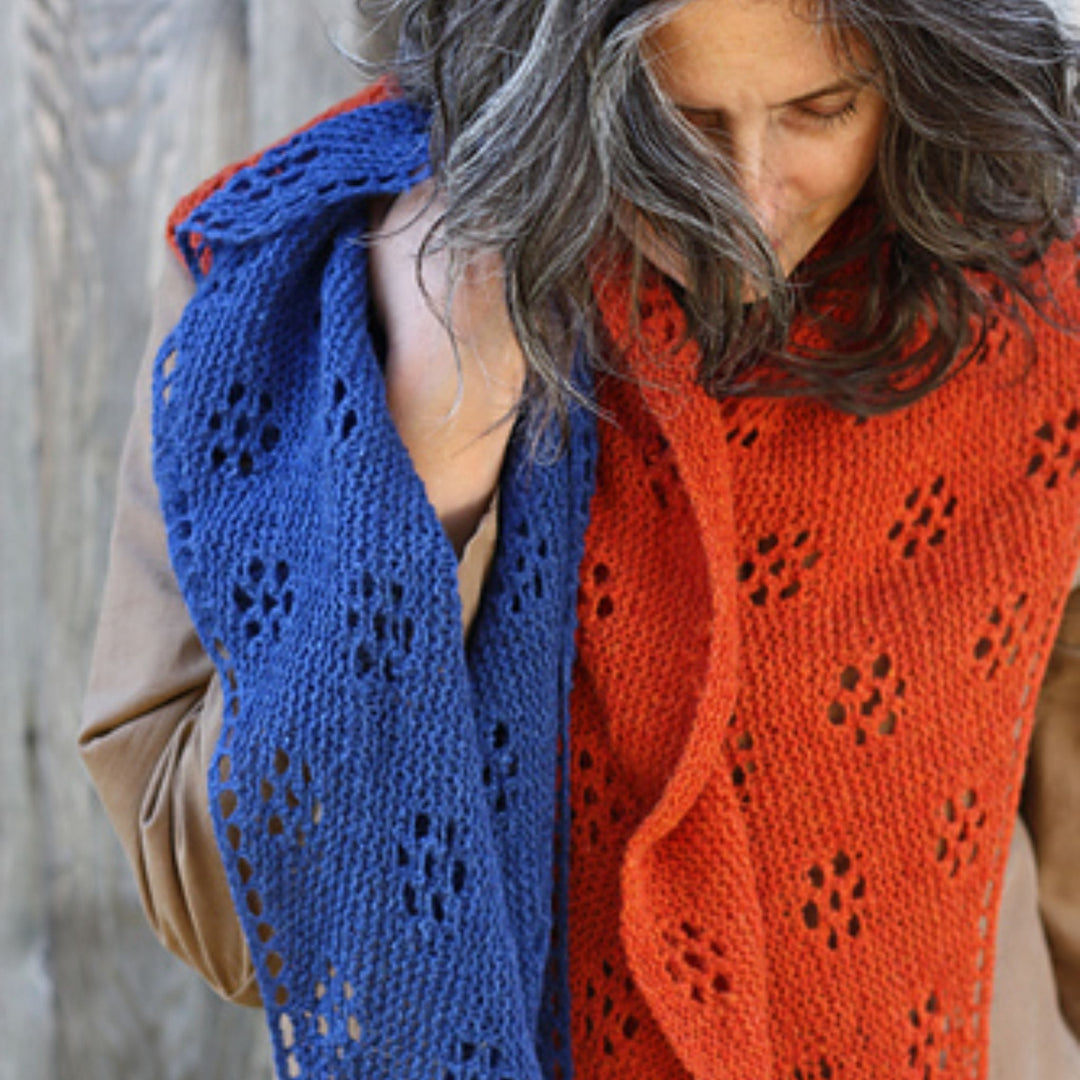 Woman wearing a blue and red knitted shawl with a neutral background and blue shawl in hand. Shawl design is Ghost Flowers by Amy Christoffers.