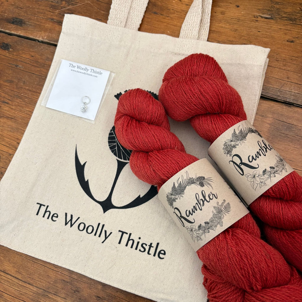 Yarn set components for Ghost Flowers Shawl by Amy Christoffers are shown at an angle and include 2 skeins of Rambler yarn in Zinnia colorway and a The Woolly Thistle Tote and Stitch marker. 