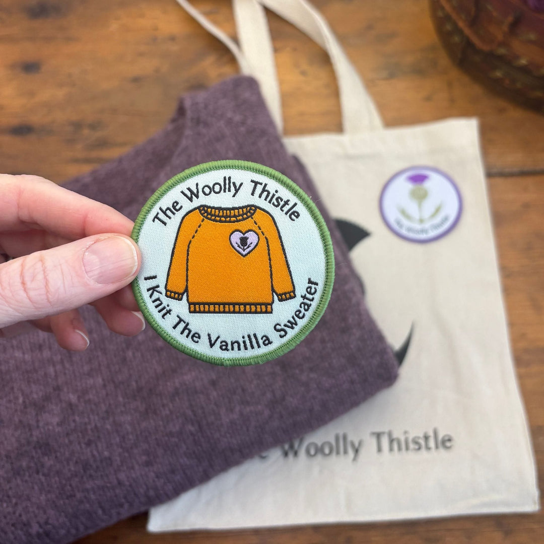 Round patch with "The Woolly Thistle I Knit the Vanilla Sweater" text and a drawing of an orange sweater. The patch is held over a wooden surface with a folded mauve sweater and cream colored tote bag.