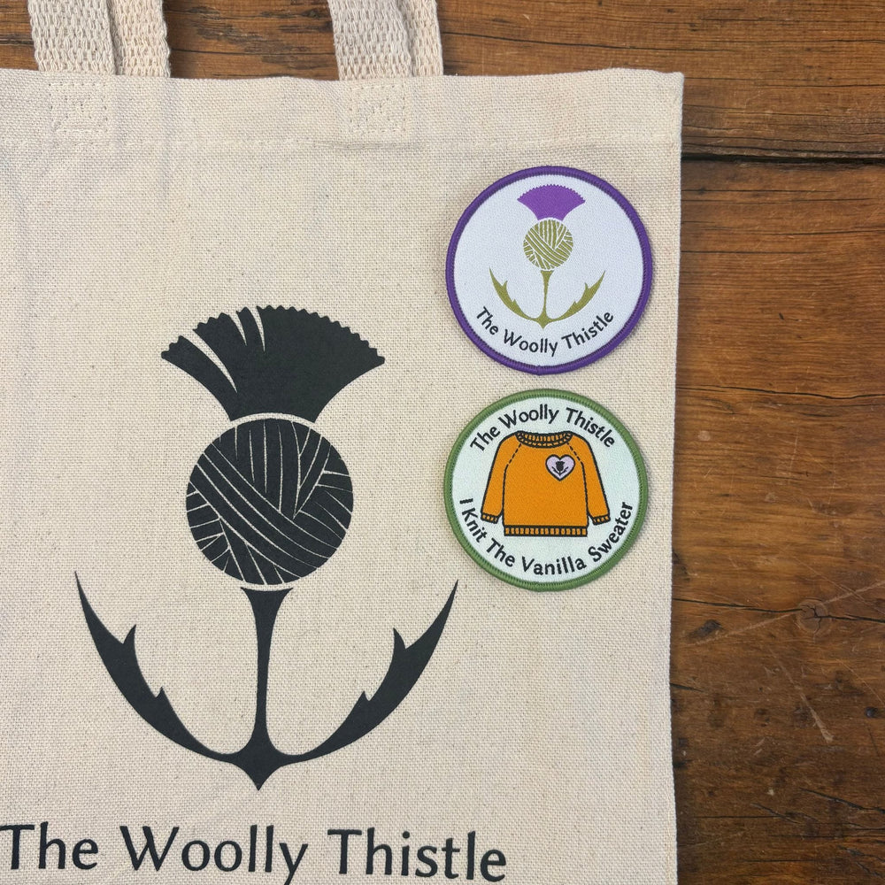 Tote bag with 'The Woolly Thistle' logo laying on a wooden surface. On the tote bag are two patches, one with The Woolly Thistle logo and one with a drawing of an orange sweater and text: "I Knit the Vanilla Sweater"
