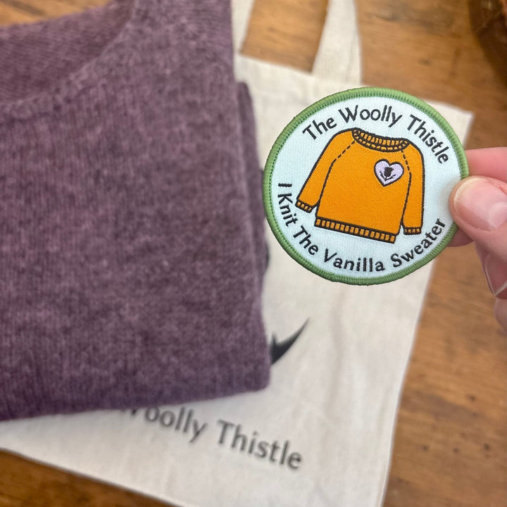 Round patch with "The Woolly Thistle I Knit the Vanilla Sweater" text and a drawing of an orange sweater. The patch is held over a wooden surface with a folded mauve sweater and cream colored tote bag.