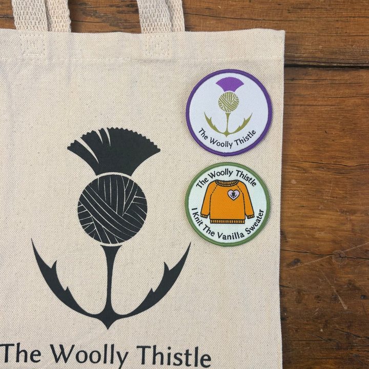 Shown on a wooden surface, a Tote bag with black and white The Woolly Thistle logo with 2 iron on patches shown in corner of bag. 'The Woolly Thistle' logo patch and "I Knit the Vanilla Sweater" patch featuring a drawing of an orange vanilla sweater. 