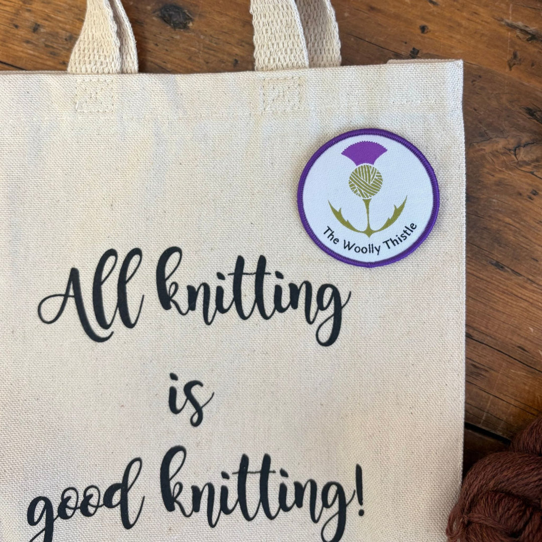 Tote bag on a wooden surface with a tote bag with 'All knitting is good knitting!' text and an iron-on patch with The Woolly Thistle logo. 