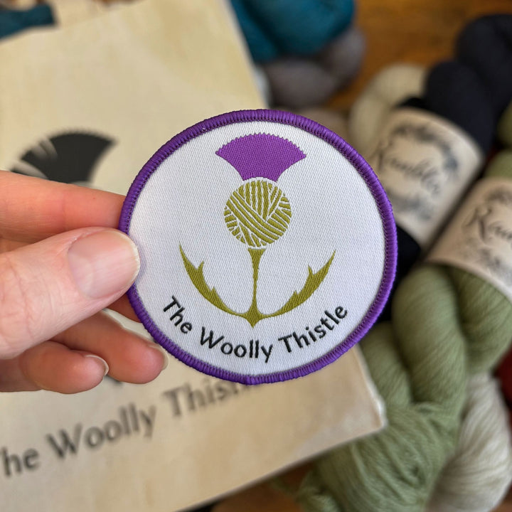 Round patch with 'The Woolly Thistle' logo held over a logo tote bag and skeins of Rambler yarn in the background.