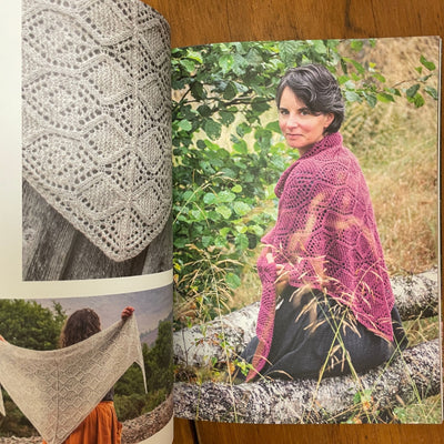 Perspectives book edited by Julie Rutter & Emily K. Williams. Pages open to photo of the Feshie Shawl, one knit in fuschia and one in grey, draped over a woman turned towards camera while sitting on a log outside.