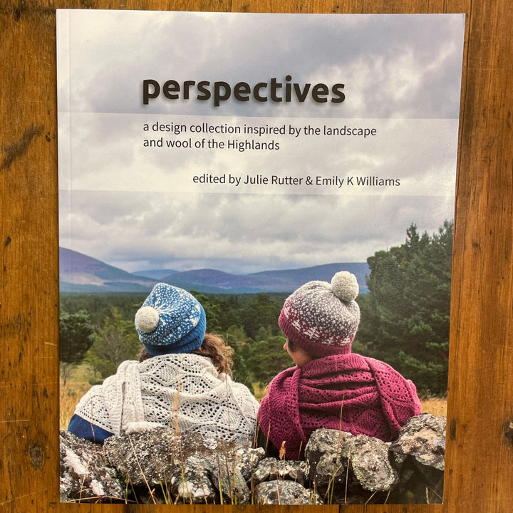 Perspectives book edited by Julie Rutter & Emily K. Williams lying on wooden table. Book cover photos shows view from behind of two women sitting in a field wearing handknit hats.