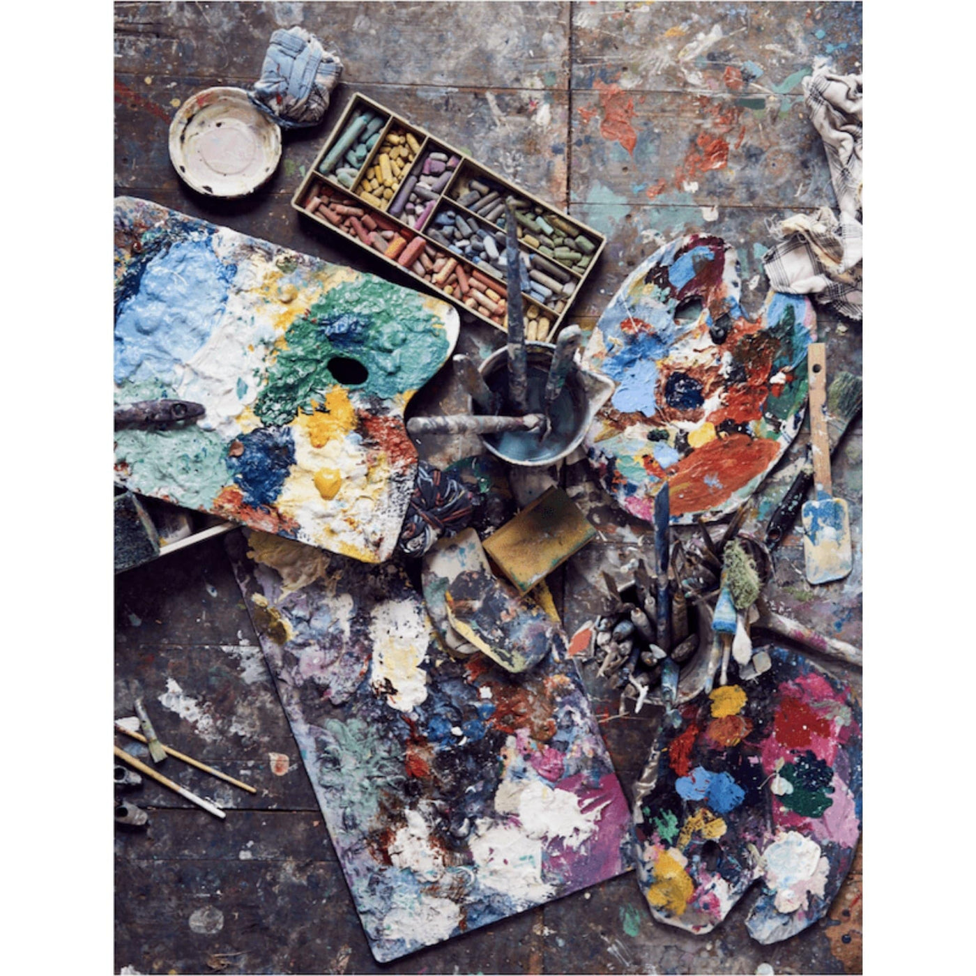 A photo of artists paint palettes on the floor along with tools, bruses and pastels.