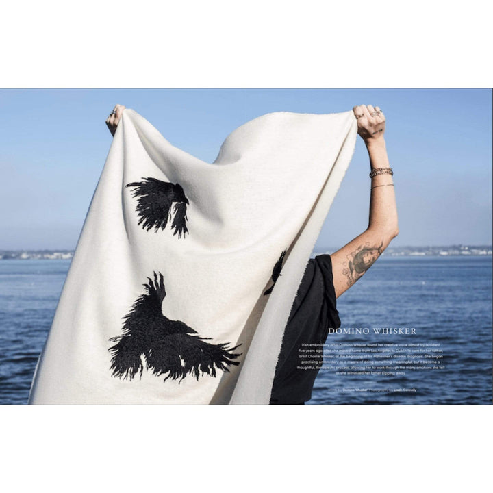 A white person standing by a lake holds up a piece of fabric with flying crows printed on it.