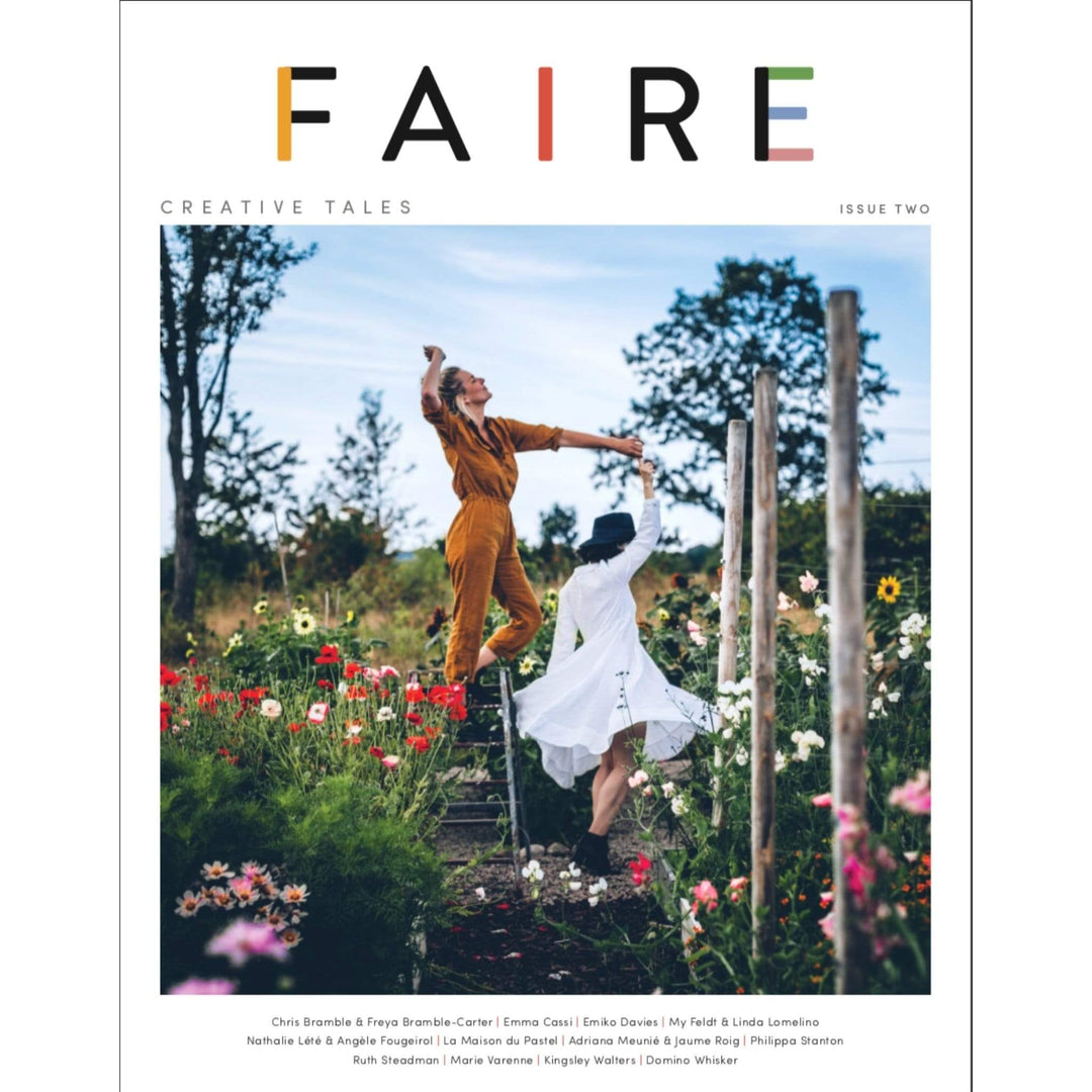 The cover of Faire Issue two features two white women dancing in a flower garden on a sunny day.