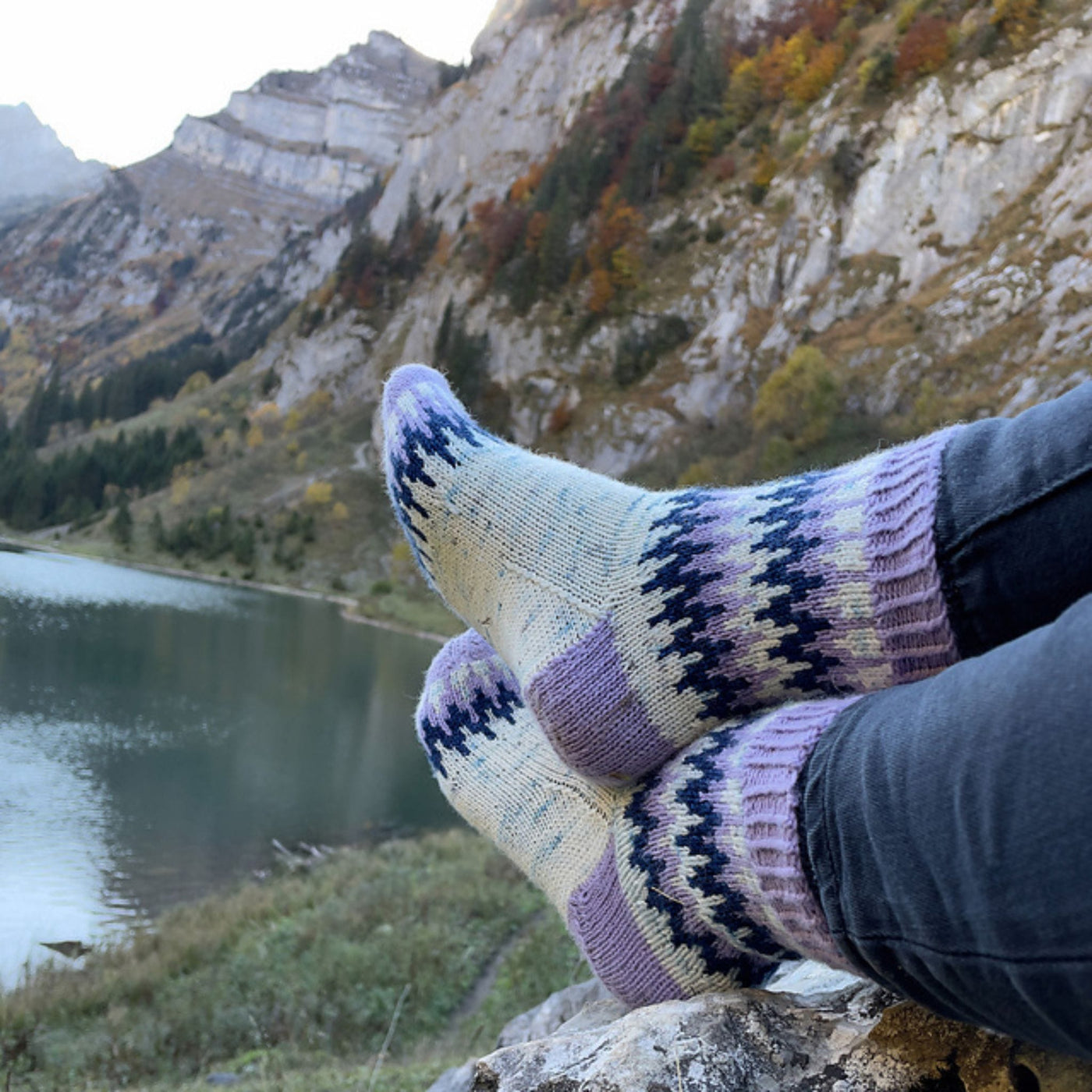 Two feet with crossed ankles near a mountain lake wearing white socks with lavender and navy colorwork