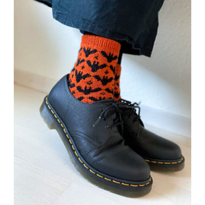 Two feet wearing black shoes on a white floor with orange socks with a black bat motif