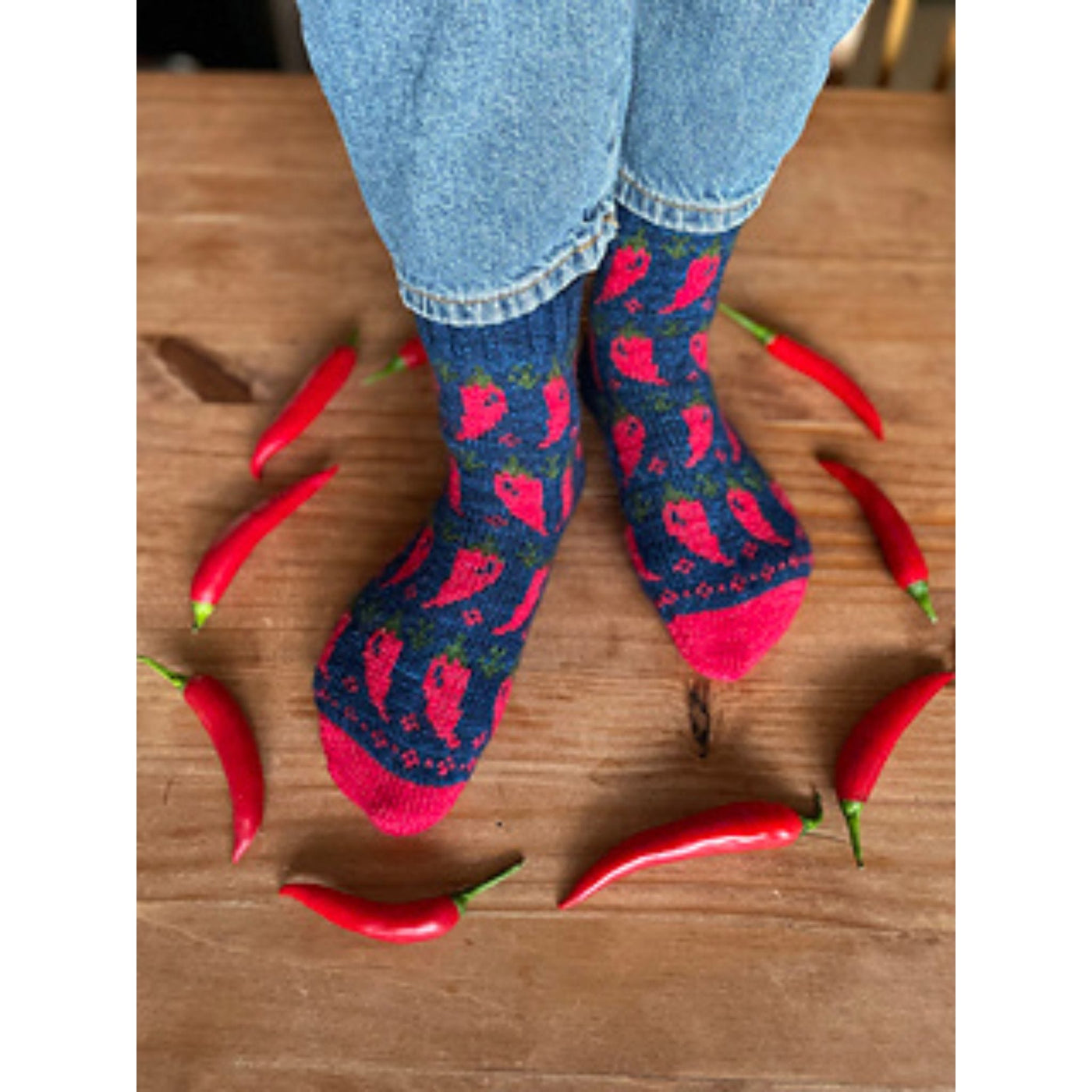 Two feet standing on a table surrounded by chile peppers wearing blue socks with a red and green chile pepper motif