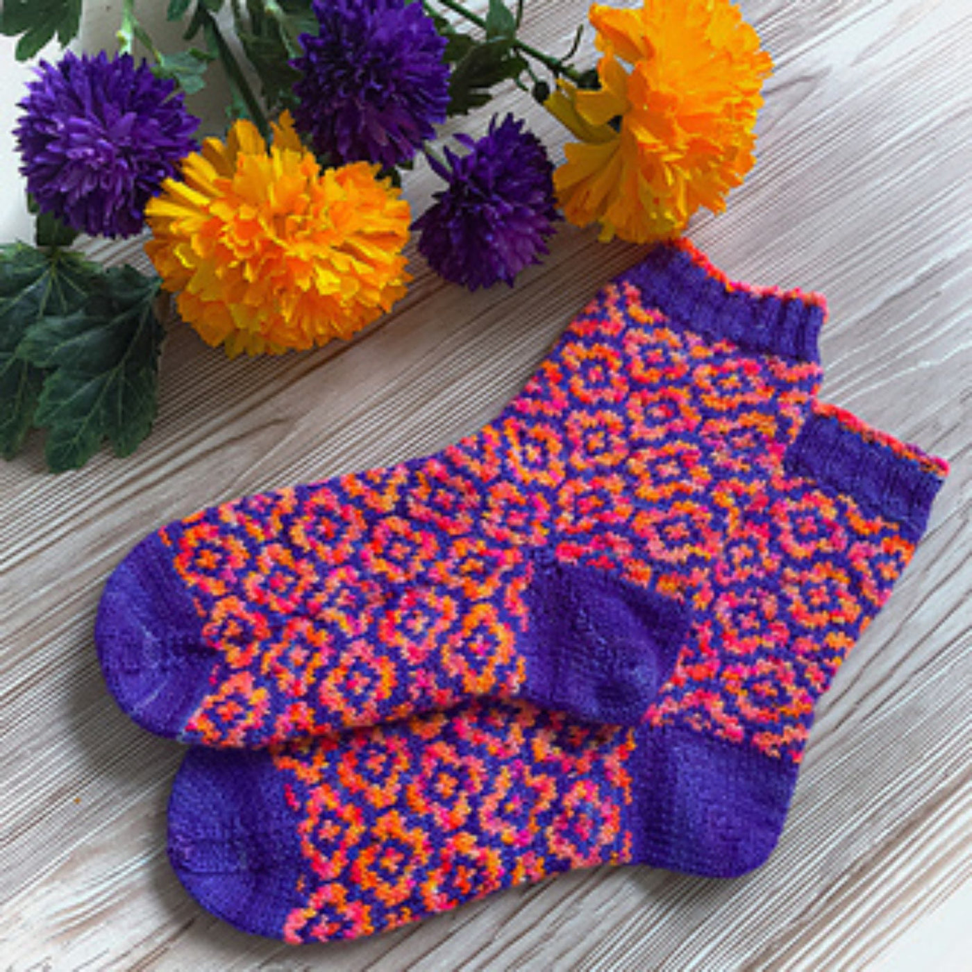 Two socks on a grey wood floor near purple and orange flowers, with purple cuffs, heels and toes and the purple, pink, and orange flower motif