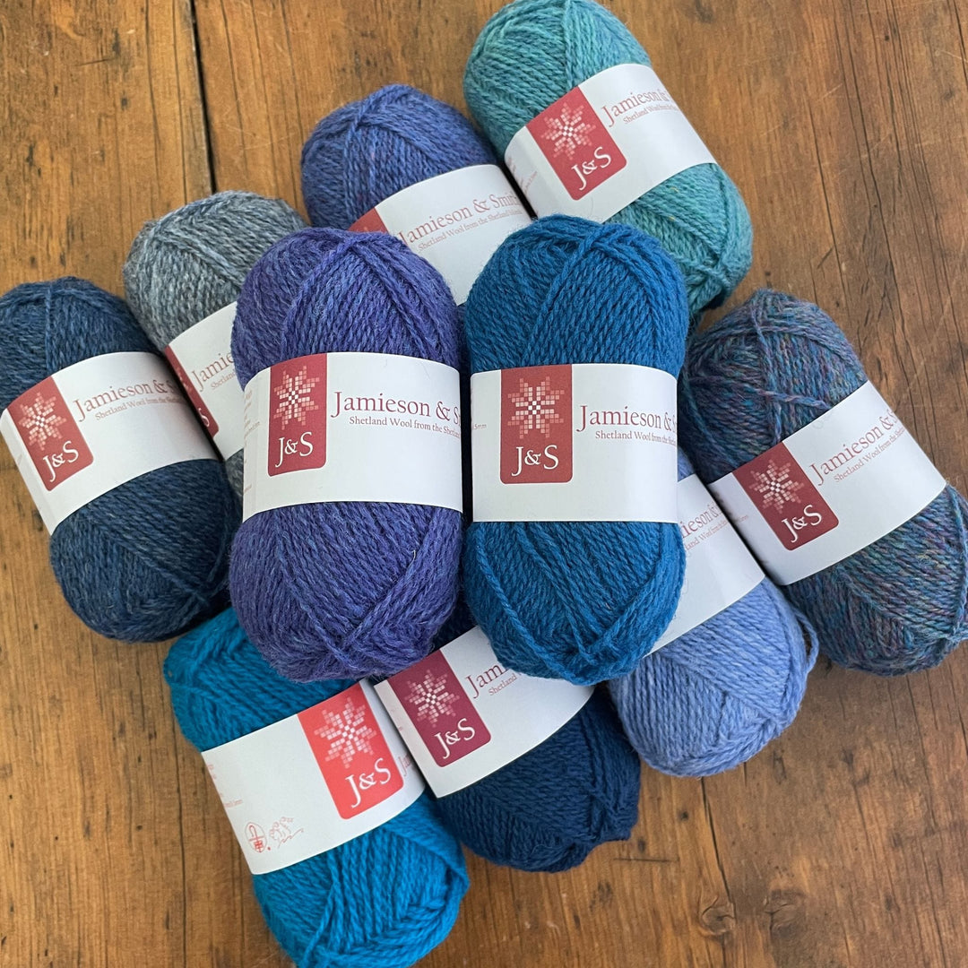Ten balls of Jamieson & Smith 2ply Jumper Weight yarn, a light fingering weight, in shades of blue, on a wooden table
