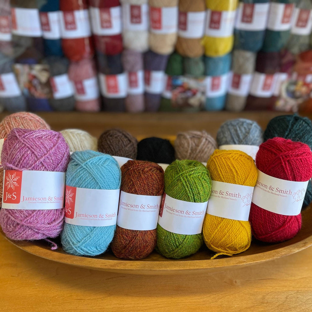 13 balls of colorful J&S yarn in a basket.  In the background grab bags of J&S yarn in many different colors. 