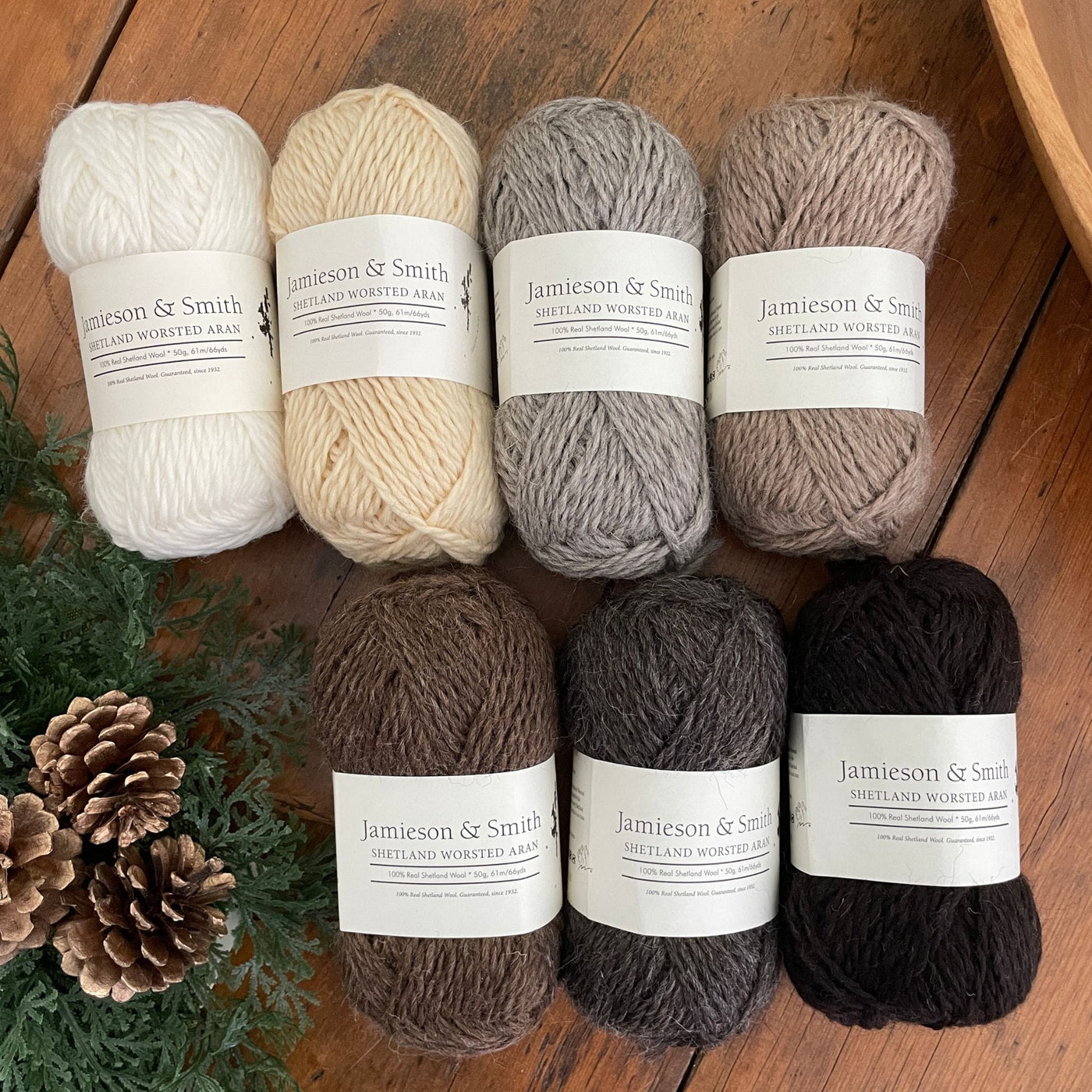 Jamieson & Smith Aran Worsted Yarn in natural shades arranged in two rows on wooden table with greenery and pinecones.
