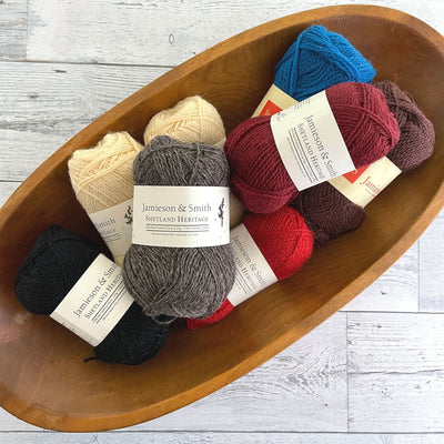 Jamieson & Smith Shetland Heritage Yarn in variety of colors. Neutrals, reds, and blue yarn in oval-shaped wooden bowl.