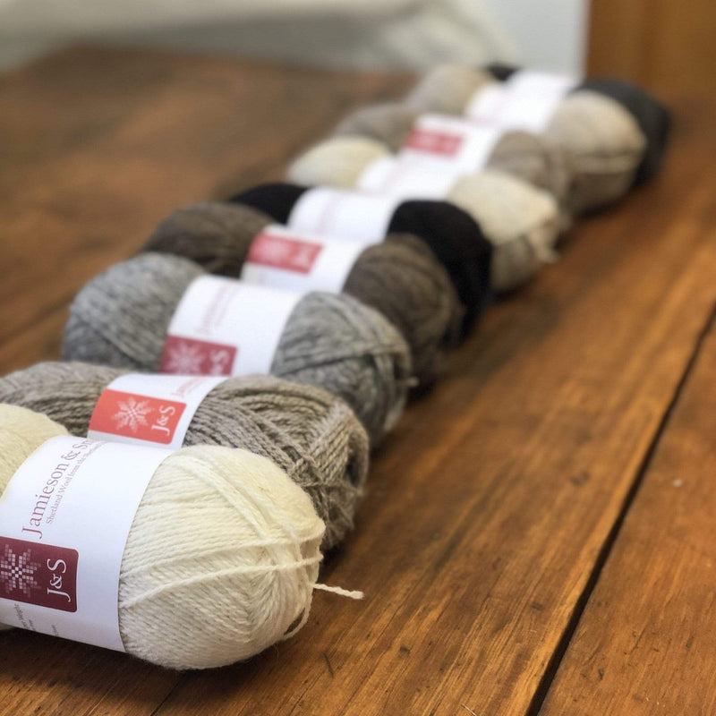 An array of balls of Jamieson & Smith Shetland Supreme, a fingering weight wool yarn, in various colors, on a wooden table 