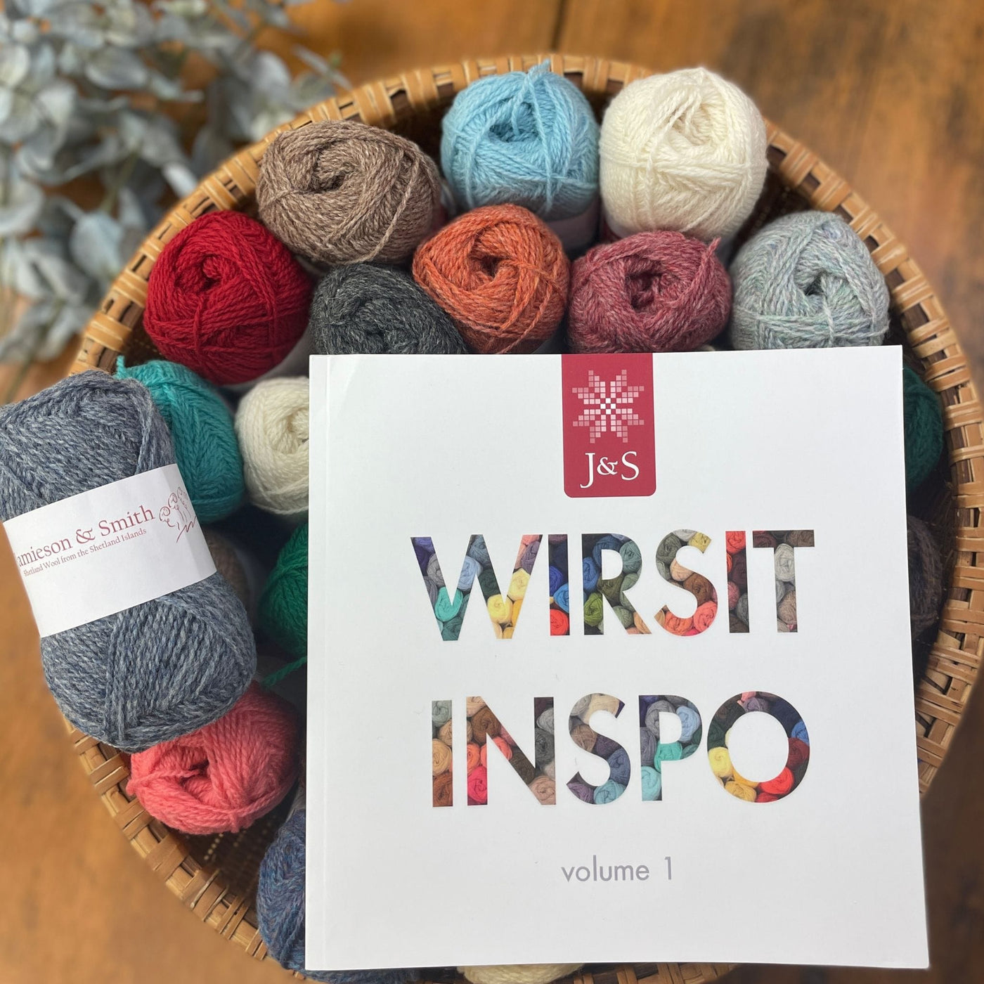 Wirsit Inspo Volume 1 from Jamieson & Smith. Cover of book is shown on top of a basket of Jamieson & Smith 2ply Jumper Weight yarn. 