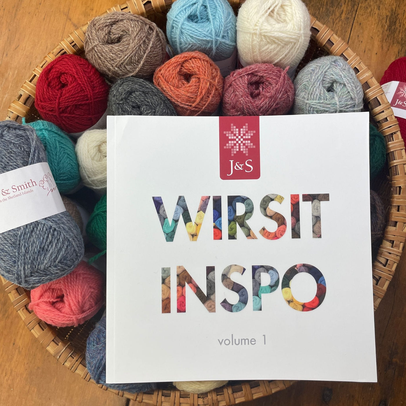 Wirsit Inspo Volume 1 from Jamieson & Smith. Cover of book is shown on top of a basket of Jamieson & Smith 2ply Jumper Weight yarn. 