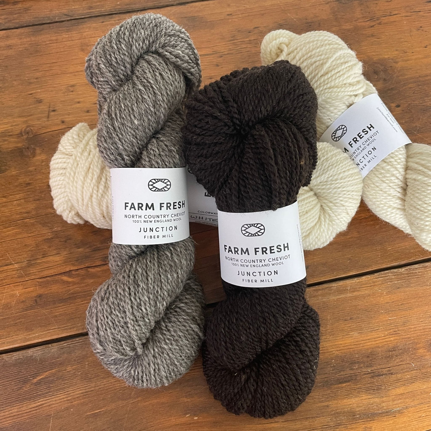 Four skeins of Junction Fiber Mill Farm Fresh, a DK weight yarn, in three natural colorways, on a wooden table