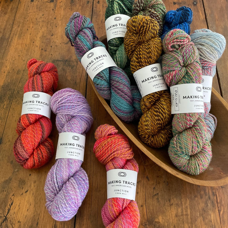 Ten skeins of Junction Fiber Mill Making Tracks yarn, a DK weight, barberpole yarn, in various colors, on a wooden table and in a wooden bowl