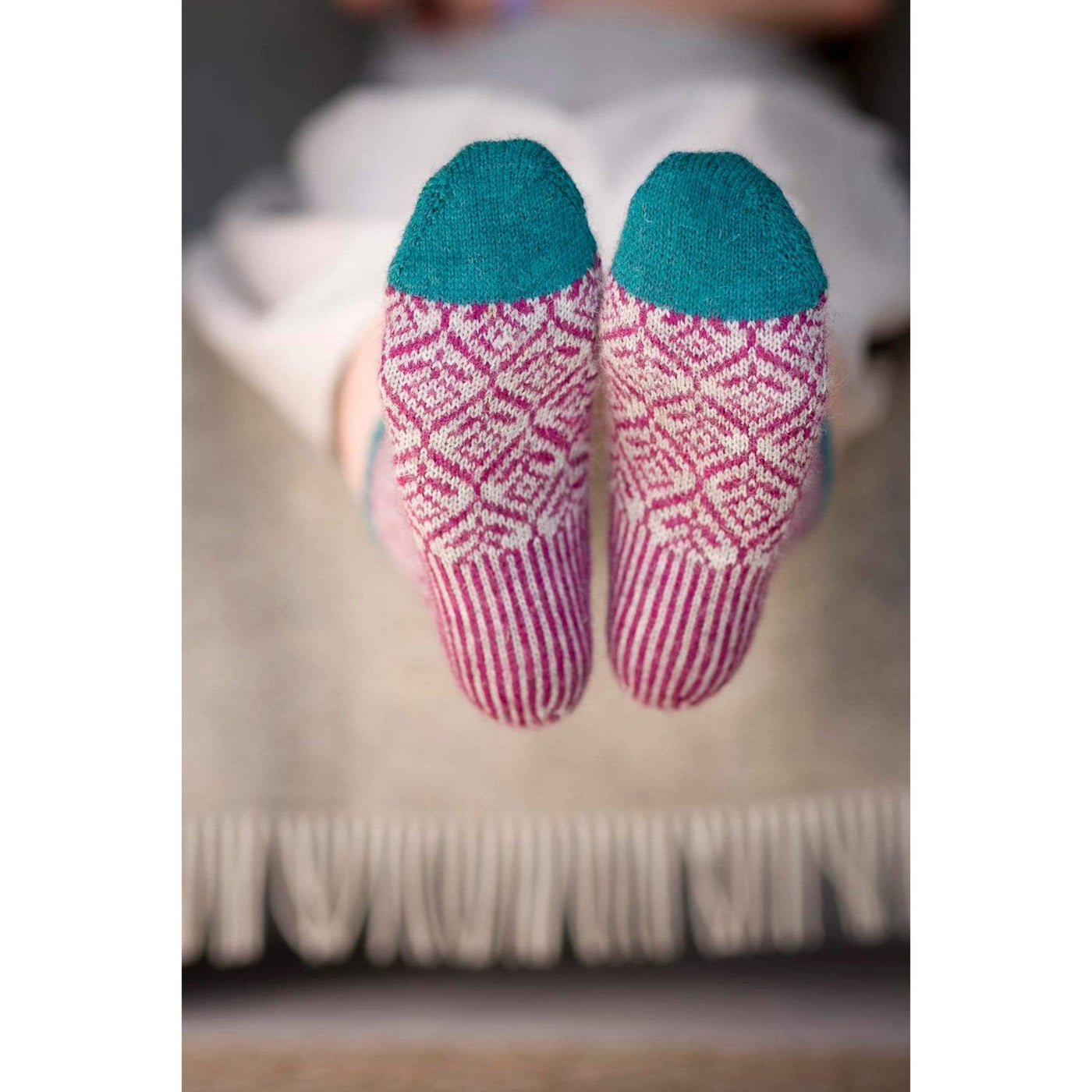 The Woolly Thistle Bluestockings by Kate Davies & Nicole Pohl image of woman sitting down showing soles of pink and beige  patterned with color blue on toes area  knitted socks 