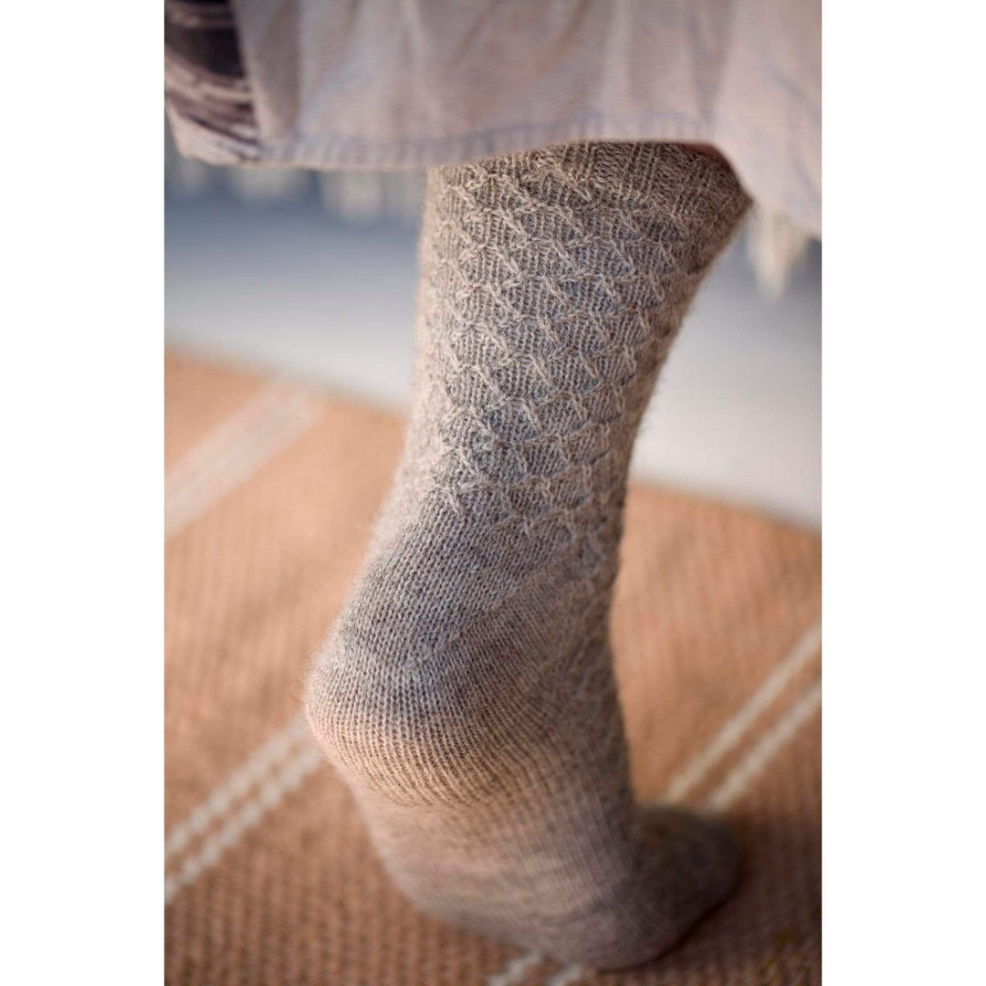 The Woolly Thistle Bluestockings by Kate Davies & Nicole Pohl featuring woman wearing beige knitted socks with a dress