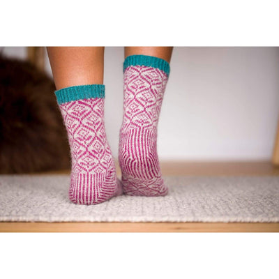 The Woolly Thistle Bluestockings by Kate Davies & Nicole Pohl featuring woman wearing pink and beige knitted socks with blue stripes around ankle area