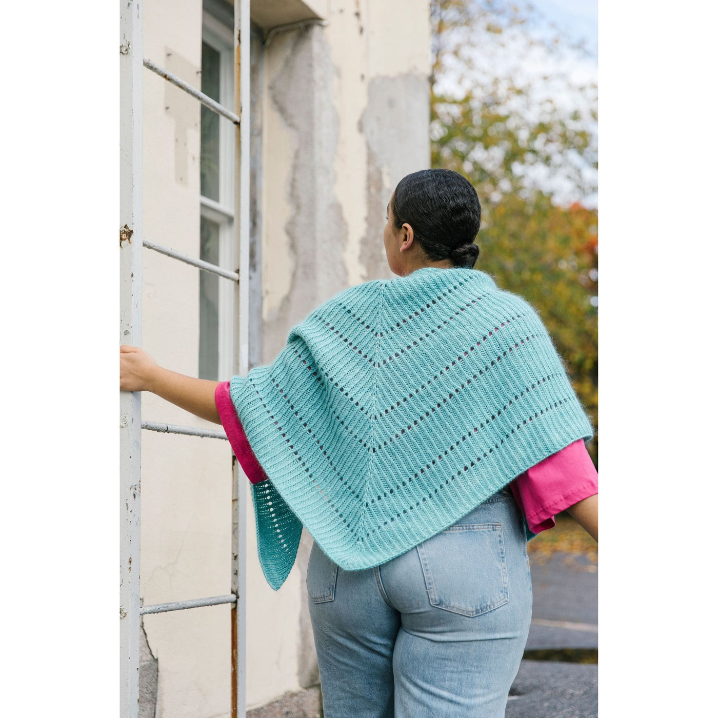 52 Weeks of Easy Knits page shows model wearing one of the knit designs in the book. View of woman from behind. Woman in next a building wearing jeans, a pink shirt, and light blue shawl draped down her back. 