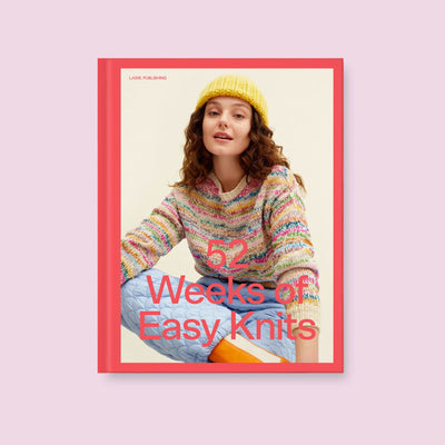 Cover of 52 Weeks of Easy Knits by Laine shows woman wearing multicolor sweater and yellow knit hat. Book background is pink and book is set on light pink background. 