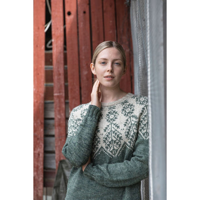 Knitted Kalevala book by Jenna Kostet from Laine Publishing. A design from book is shown on woman standing in front of boards wearing a green and beige colorwork sweater.