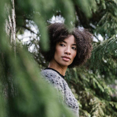 Model photgraphed through trees wearing a gray knit sweater with bobbles.
