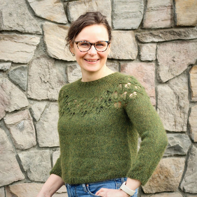 Woman wearing Love Note Sweater in Green with lacework yoke, standing in front of stone wall.