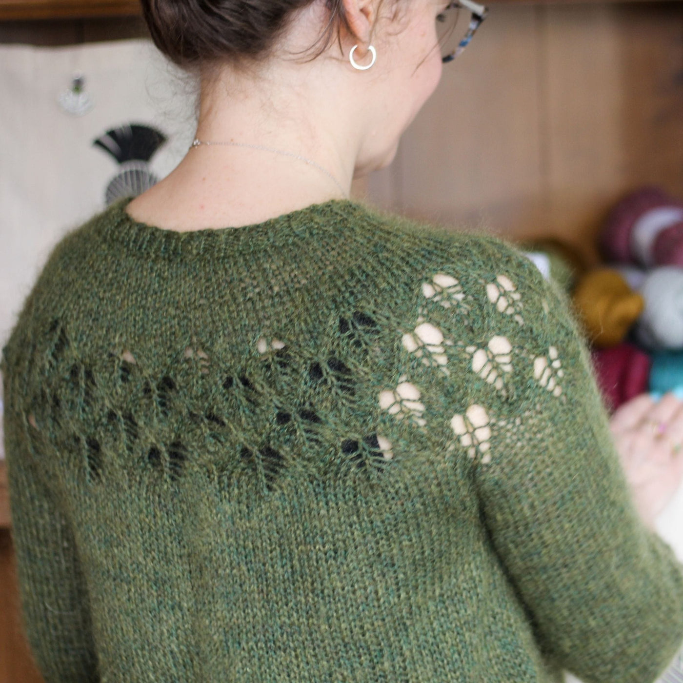 Back view of woman wearing Love Note Sweater in Green with lacework yoke, standing in front of shelf of yarn.
