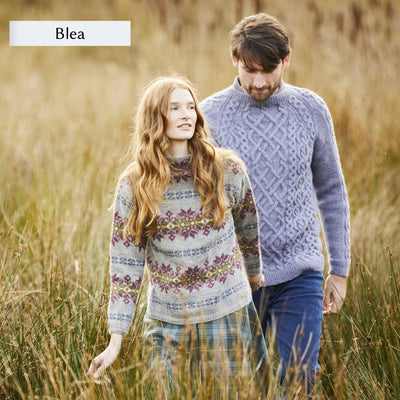 Two models wearing designs from Westmorland book by Marie Wallin. Models are walking in grassy field. Woman is wearing an allover colorwork sweater and man is wearing a textured grey sweater.