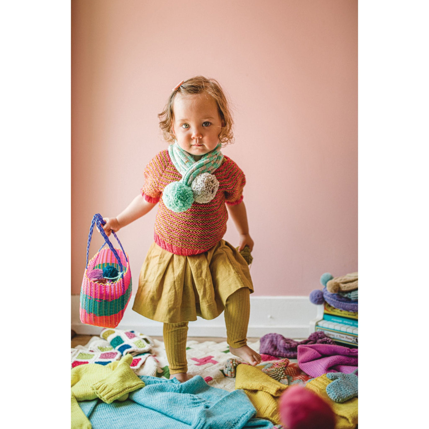 Child surrounded by knit items wearing pink, colorful sweater and pom pom scarf which are designs from Mini Pom Magazine. 
