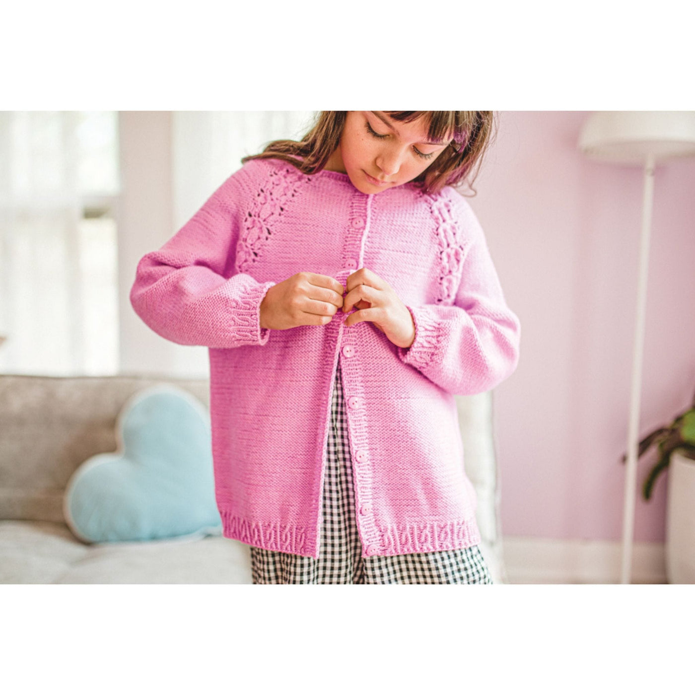 Child wearing pink cardigan looking down while buttoning the front.  Cardigan is a design from Mini Pom Magazine. 