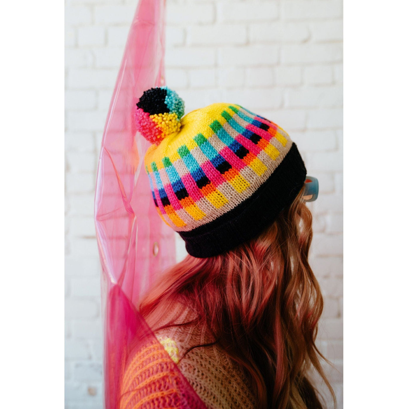 A woman wearing a bright and colorful knit hat featuring bands of bright colors and topped with a colorful pompom.